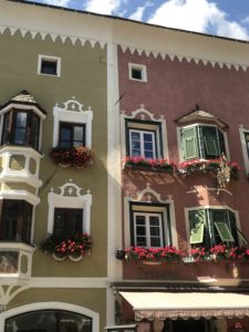 Ein grünes Haus und ein rosanes Haus, jeweils mit weißen Fensterumrandungen, Stuck und Blumen an den Fenstern