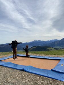 Die Kinder hüpfen auf einem in den Boden eingelassenem Trampolin vor dem Bergpanorama im Hintergrund