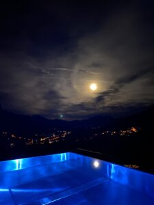 Ein Blick vom Infinityhotelppl in den Nachthimmel bei Vollmond. Dahinter sieht man schwarzes Bergpanorama und die Orte im Lichterglanz